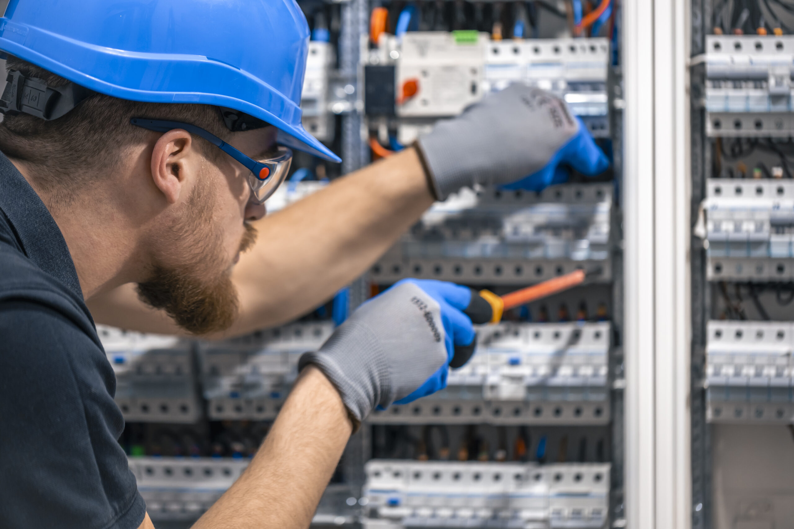 A male electrician works in a switchboard with an electrical connecting cable. Electrician with screwdriver tightens electrically operated switching equipment in fuse box.